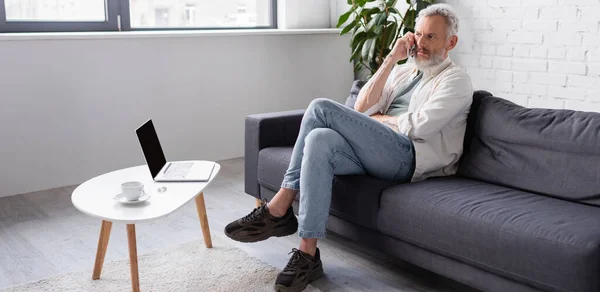 Homem barbudo falando no smartphone enquanto sentado no sofá perto do laptop com tela em branco, banner — Stock Photo