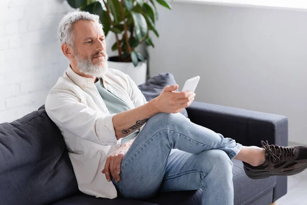 Bärtiger Mann sendet Nachrichten auf dem Smartphone, während er auf der Couch sitzt — Stockfoto