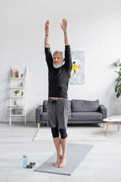 Vollbärtiger und tätowierter Mann beim Training auf Fitnessmatte in der Nähe von Sportflasche und Hanteln im Wohnzimmer — Stockfoto