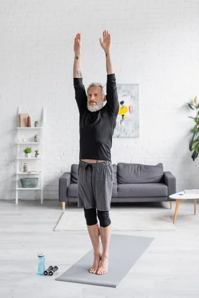 Homme barbu et tatoué faisant de l'exercice sur un tapis de fitness près d'une bouteille de sport et d'haltères dans le salon — Photo de stock