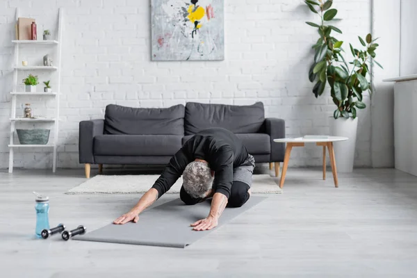 Mann mit grauen Haaren streckt sich auf Fitnessmatte neben Hanteln im Wohnzimmer, Banner — Stockfoto