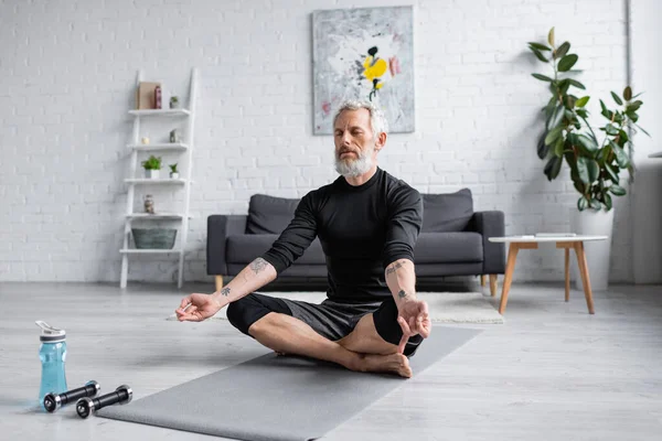 Hombre con pelo gris meditando en esterilla de yoga cerca de pesas en la sala de estar, pancarta - foto de stock