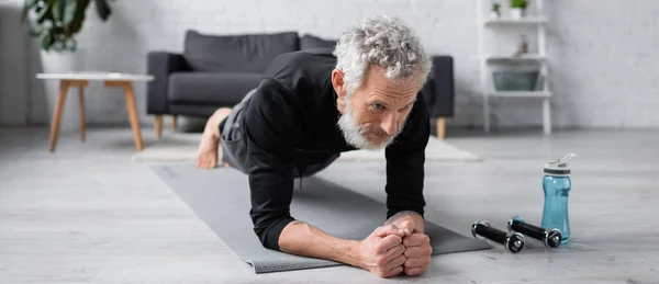 Homme barbu faisant planche sur tapis de fitness près des haltères dans le salon, bannière — Photo de stock