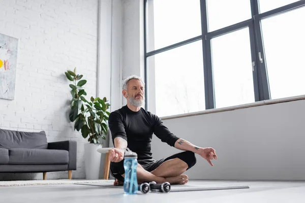 Bearded man with grey hair meditating on yoga mat near dumbbells in living room — Stock Photo