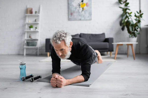 Hombre barbudo con pelo gris haciendo tablón en la alfombra de fitness cerca de pesas en la sala de estar - foto de stock
