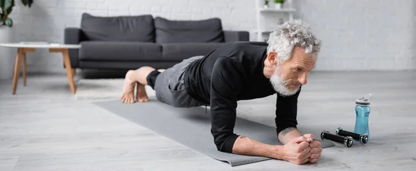 Hombre descalzo con el pelo gris haciendo tablón en la estera de fitness cerca de pesas en la sala de estar, pancarta - foto de stock