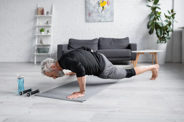 Hombre descalzo con el pelo gris haciendo ejercicio en la alfombra de fitness cerca de pesas en la sala de estar - foto de stock