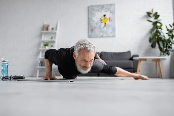 Strong man with grey hair working out on fitness mat near dumbbells in living room — Stock Photo