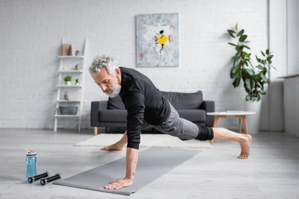 Hombre deportivo con el pelo gris haciendo ejercicio en la alfombra de fitness cerca de pesas en la sala de estar - foto de stock