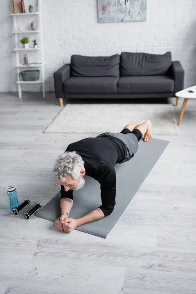 Visão de alto ângulo do homem com cabelos grisalhos exercitando-se no tapete de fitness perto de halteres na sala de estar — Fotografia de Stock