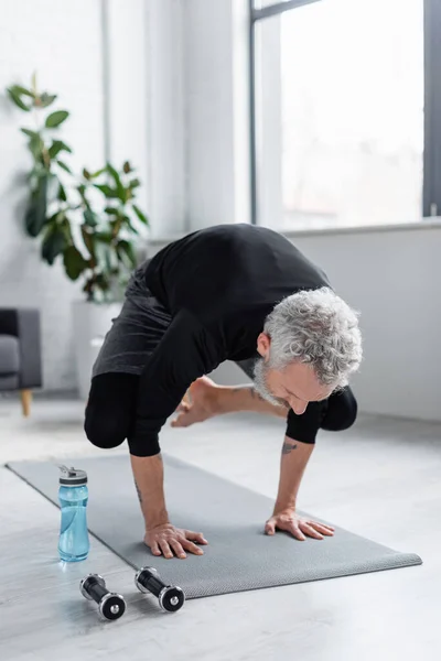 Homme fort avec les cheveux gris exercice sur tapis de fitness près des haltères et bouteille de sport dans le salon — Photo de stock