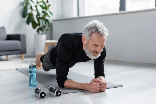 Homem forte com cabelos grisalhos fazendo prancha no tapete de fitness perto de halteres e garrafa de esportes na sala de estar — Fotografia de Stock