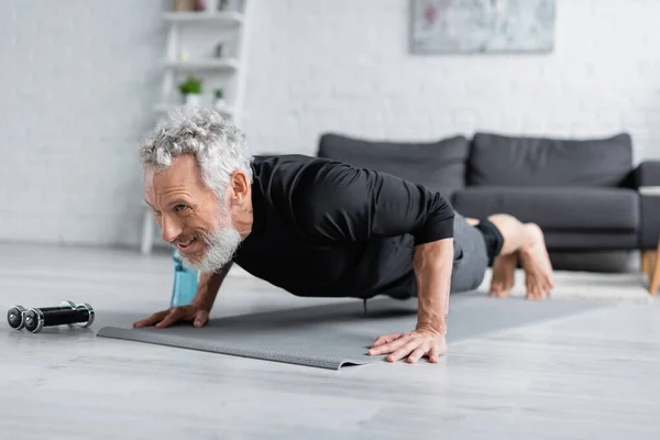 Hombre fuerte haciendo tablón en la estera de fitness cerca de pesas en la sala de estar - foto de stock