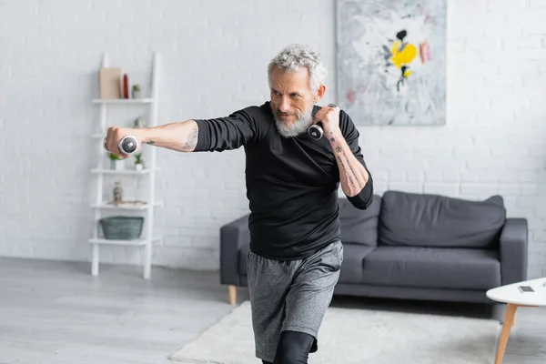 Tattooed and bearded man working out with dumbbells in living room — Stock Photo