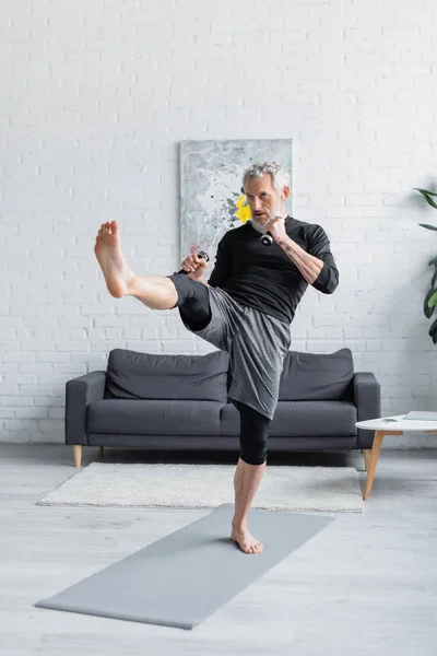 Vollbärtiger Mann, der mit Kurzhanteln trainiert, während er im Wohnzimmer kickt — Stockfoto