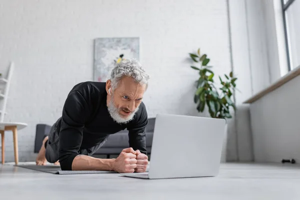 Homem barbudo em fones de ouvido sem fio fazendo prancha no tapete de fitness perto de laptop na sala de estar — Stock Photo