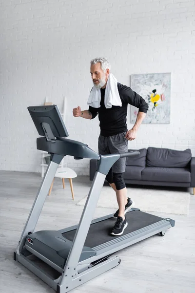 Full length of bearded man with white towel jogging on treadmill at home — Stock Photo