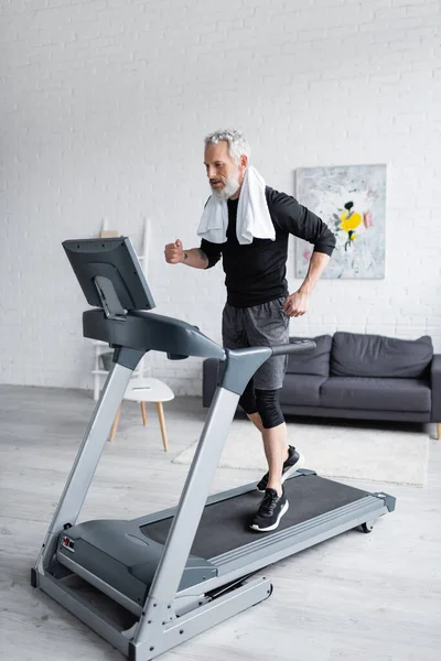 Homme barbu pleine longueur avec serviette blanche jogging sur tapis roulant à la maison — Photo de stock