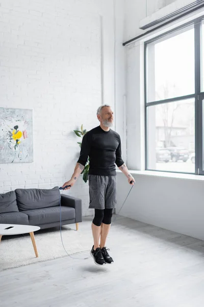 Comprimento total do homem barbudo em sportswear exercício com corda de salto na sala de estar — Fotografia de Stock