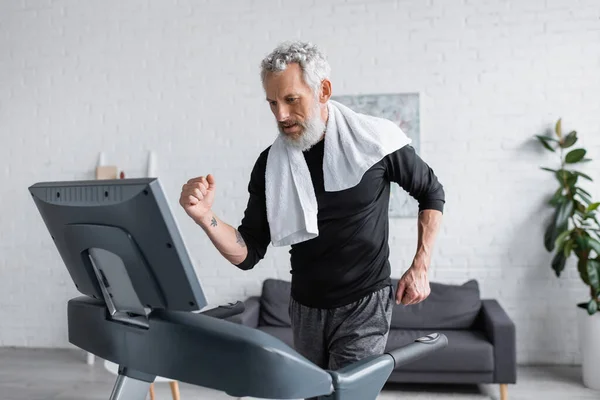 Bearded man with white towel running on treadmill at home — Stock Photo