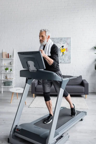 Full length of bearded man with towel running on treadmill in living room — Stock Photo