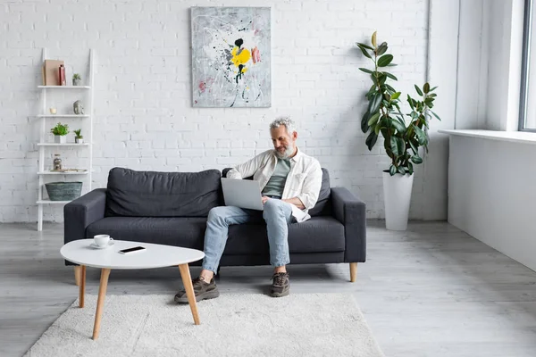 Lächelnder Mann mit Laptop sitzt auf Couch neben Tasse Kaffee und Smartphone mit leerem Bildschirm auf Couchtisch — Stockfoto