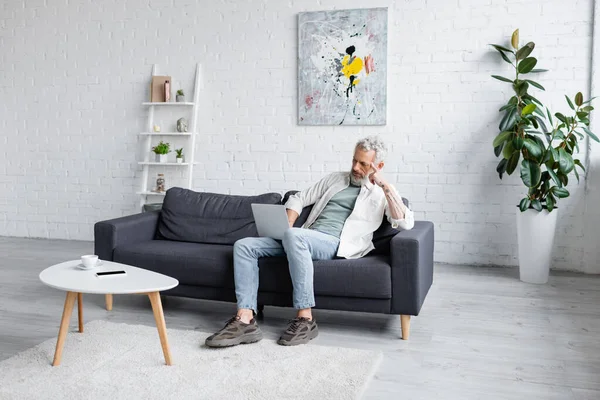 Mann mit grauen Haaren benutzt Laptop, während er auf Couch neben Tasse Kaffee und Smartphone mit leerem Bildschirm auf Couchtisch sitzt — Stockfoto