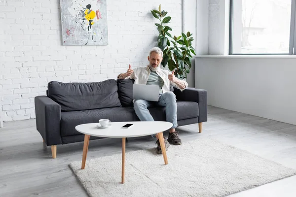 Mann mit grauen Haaren zeigt bei Videoanruf auf Laptop im Wohnzimmer Daumen hoch — Stockfoto