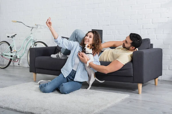Joyful young woman taking selfie with jack russell terrier and muslim boyfriend — Stock Photo