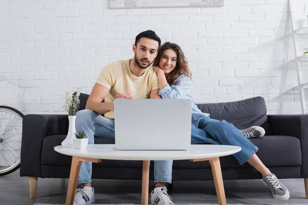Щаслива міжрасова пара дивиться фільм на ноутбуці в сучасній вітальні — стокове фото