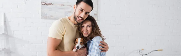 Alegre pareja multiétnica abrazando con jack russell terrier en sala de estar moderna, pancarta - foto de stock