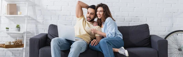 Mujer feliz apuntando a la computadora portátil cerca de novio musulmán, bandera - foto de stock