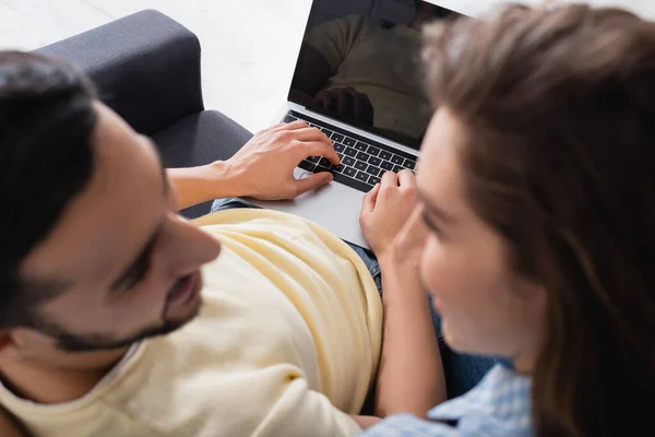 Laptop with blank screen near interracial couple looking at each other on blurred foreground — Stock Photo