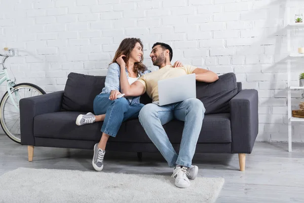 Alegre hombre musulmán y mujer feliz mirándose el uno al otro cerca de la computadora portátil en la sala de estar - foto de stock