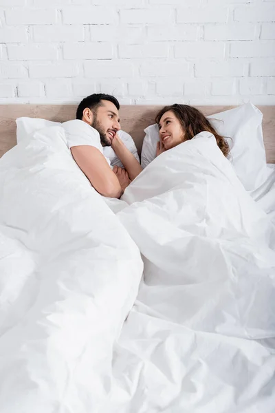 Feliz casal interracial olhando um para o outro enquanto deitado na cama — Fotografia de Stock