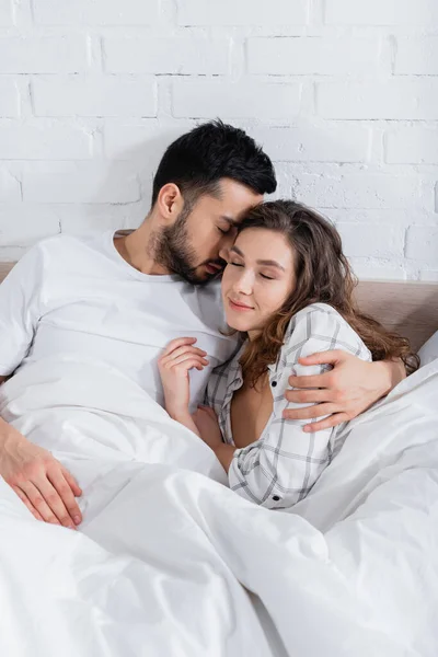 Barbudo musulmán hombre abrazando novia con los ojos cerrados en la cama - foto de stock