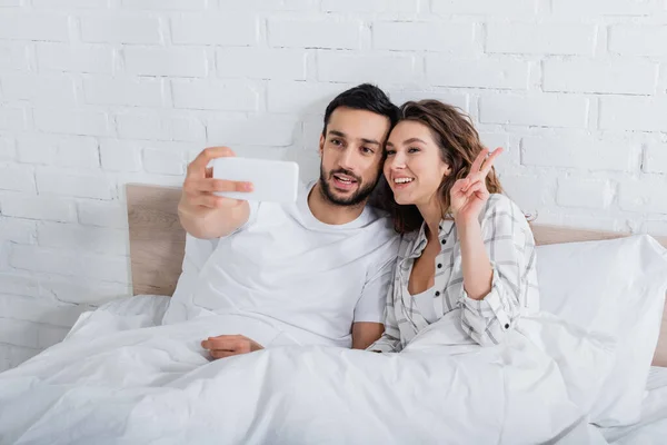 Bearded muslim man taking selfie while happy woman showing peace sign in bedroom — Stock Photo