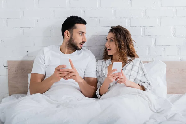 Міжрасові пари дивляться один на одного лежачи на ліжку і використовуючи смартфони — стокове фото