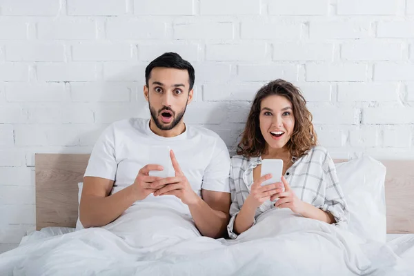 Sorprendido pareja interracial acostado en la cama y el uso de teléfonos inteligentes - foto de stock