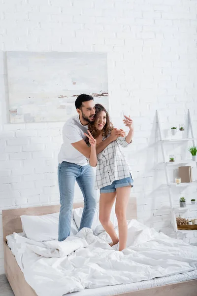 Cheerful woman standing on bed and taking selfie with mulsim boyfriend while showing peace sign — Stock Photo