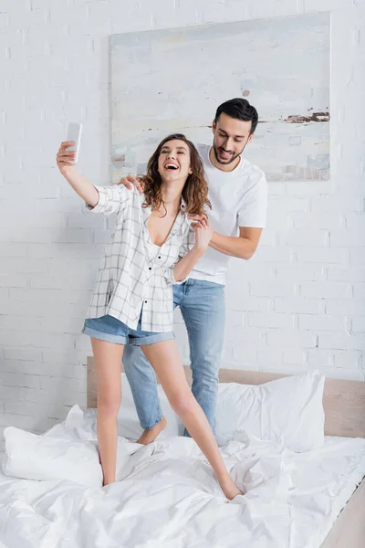 Excited woman standing on bed and taking selfie with mulsim boyfriend — Stock Photo