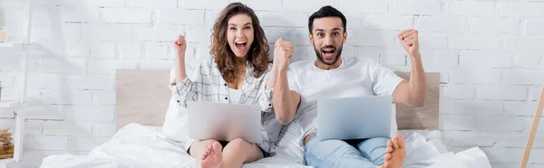 Excited multiethnic freelancers lying on bed with laptops, banner — Stock Photo