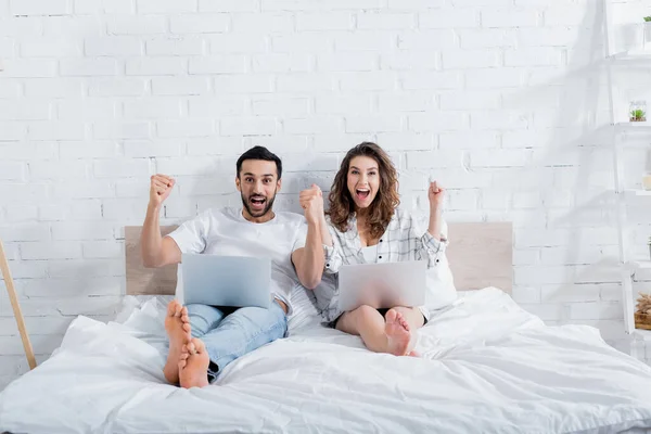 Happy multiethnic freelancers lying on bed with laptops — Stock Photo