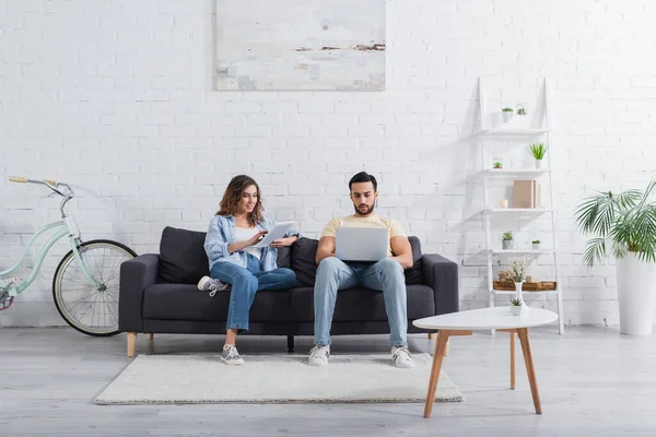 Muslim freelancer using laptop near girlfriend with digital tablet on couch — Stock Photo