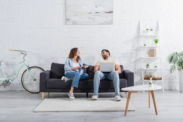 Happy muslim freelancer using laptop near girlfriend with digital tablet on couch — Stock Photo