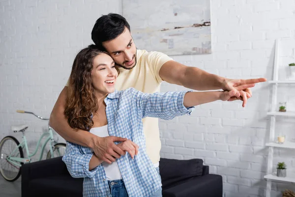 Homem multiétnico feliz e mulher abraçando e apontando com os dedos em casa — Fotografia de Stock