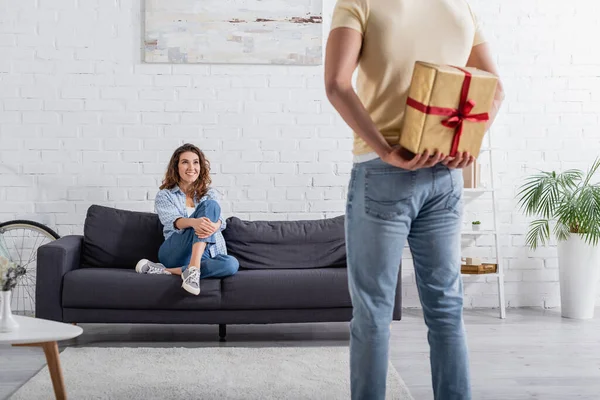 Blurred man hiding present behind back near happy woman in sofa — Stock Photo