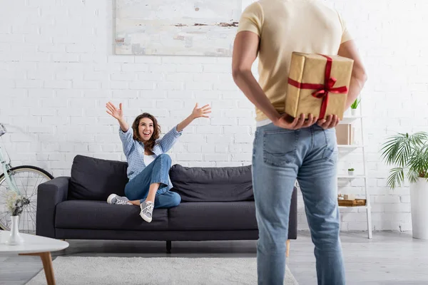 Blurred man hiding present behind back near excited woman in sofa — Stock Photo