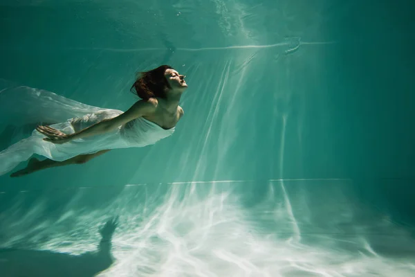 Morena joven en vestido elegante blanco nadando en la piscina - foto de stock