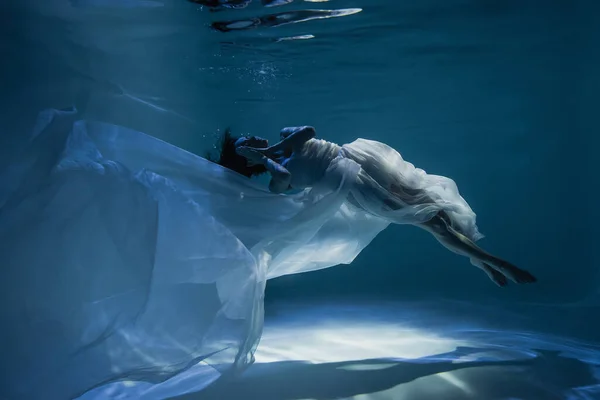 Iluminación de la mujer joven tranquila en vestido elegante blanco durmiendo mientras se nada en la piscina - foto de stock